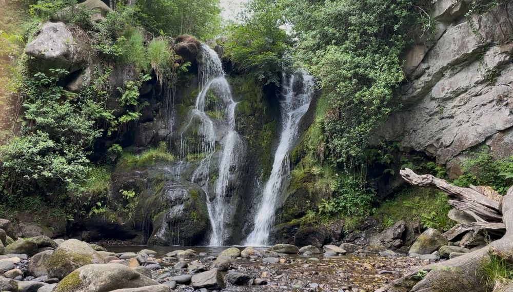 A photograph of a two-forked waterfall tumbling over mossy rocks into a shallow, pebbled pool 