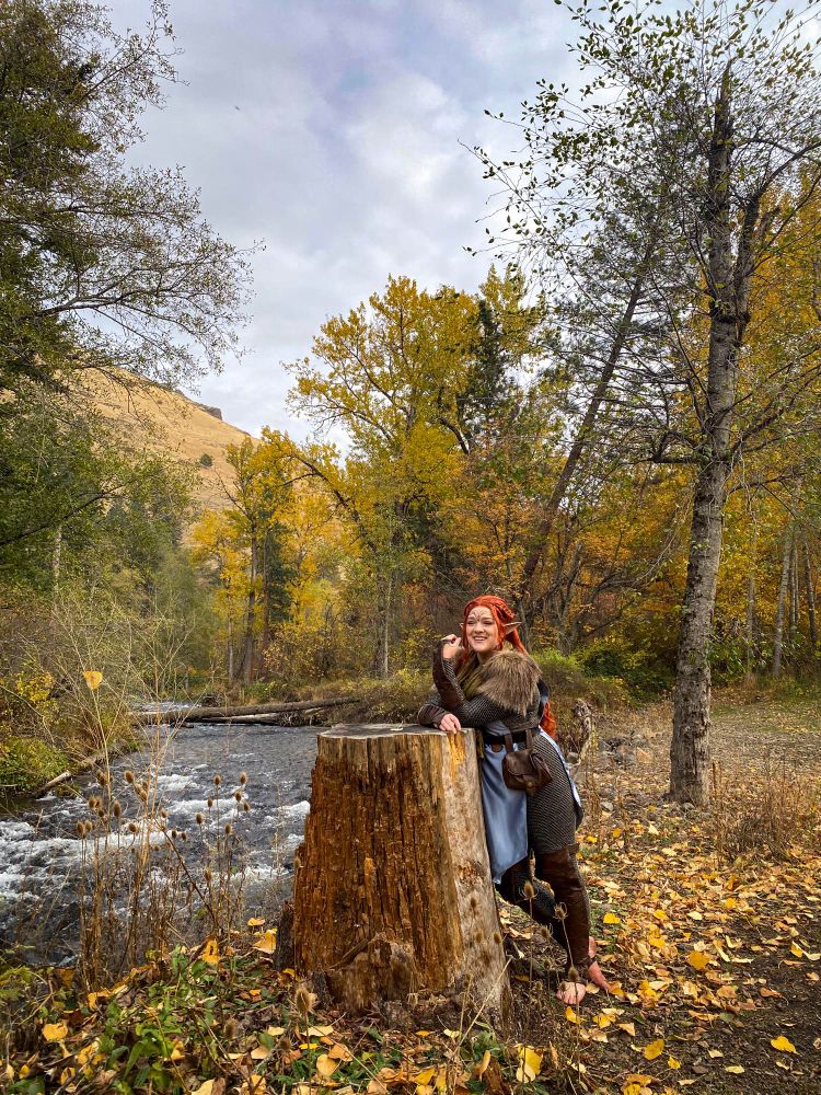 Lexi is leaning on a tree stump smiling. In the background are fall trees and a river. She is wearing a red wig, blue tunic and green scarf.