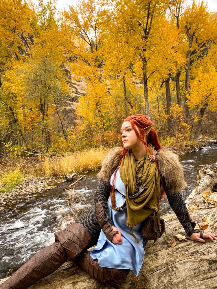 Lexi is seated on a log in front of a river and yellow trees. She is wearing a red wig, blue tunic and green scarf.