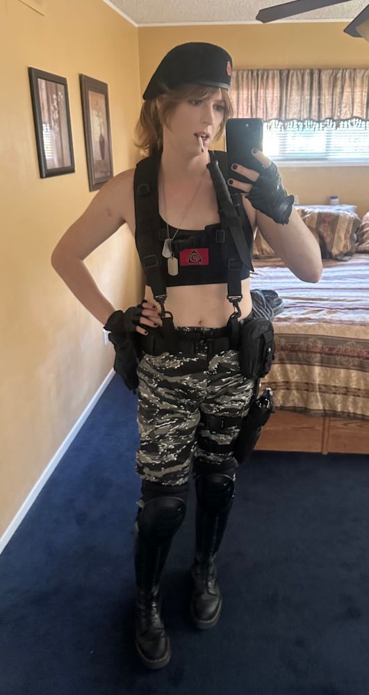 A woman dressed in a black crop top with the nod logo on it, urban tiger stripe camo pants, tall boots, kneepads, a tactical belt with shoulder straps, a drop leg holster, black fingerless gloves, and a black beret also with the NOD logo on it.