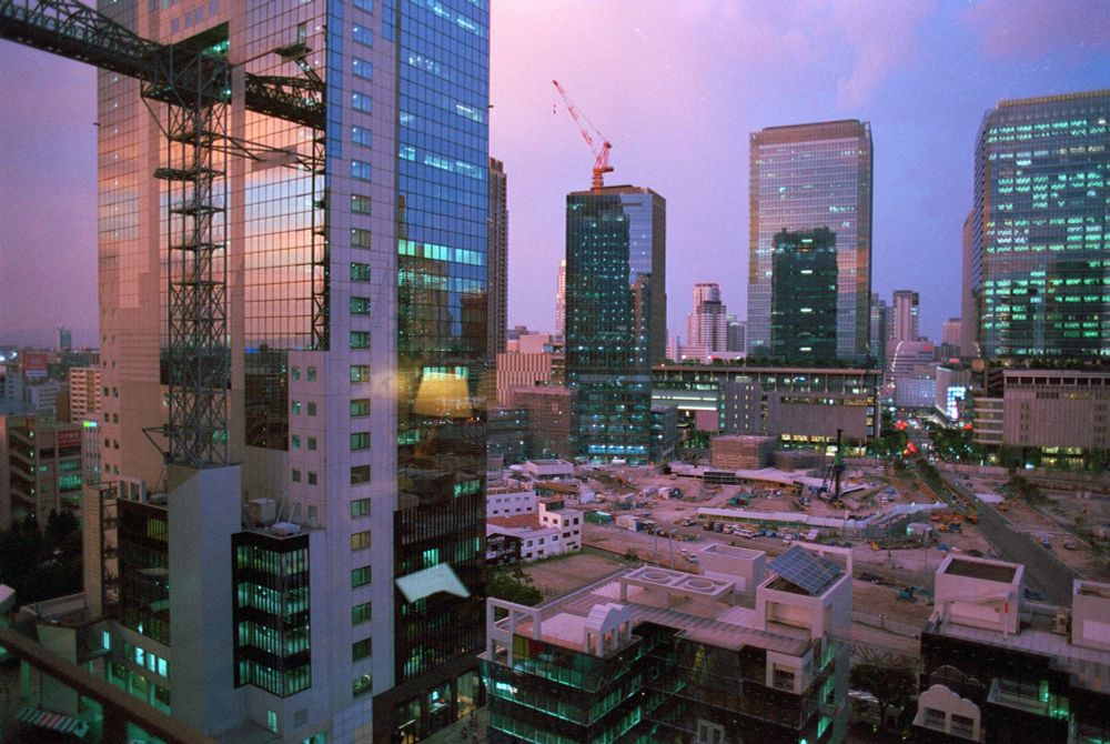 35mm photo of tall buildings of Osaka at sunset, seen through a hotel window. Construction site below. Rosy light on everything.