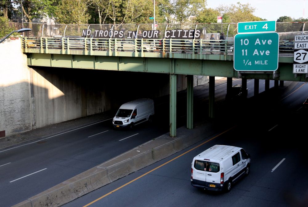 Cars drive under an overpass as Indivisible Brooklyn members hold signage over the freeway reading "NO TROOPS IN OUR CITIES"