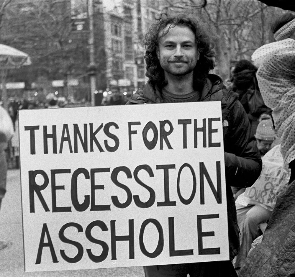 35mm B&W photo of a man smiling at the camera, holding a big sign that reads 