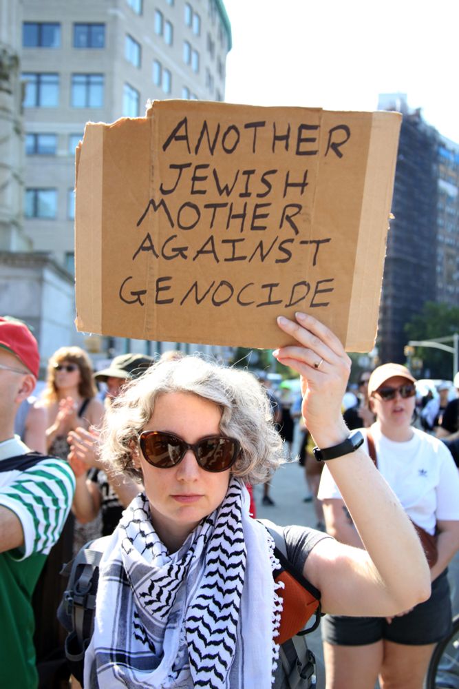 A woman with gray hair wearing a keffiyeh holds up a sign reading "ANOTHER JEWISH MOTHER AGAINST GENOCIDE"