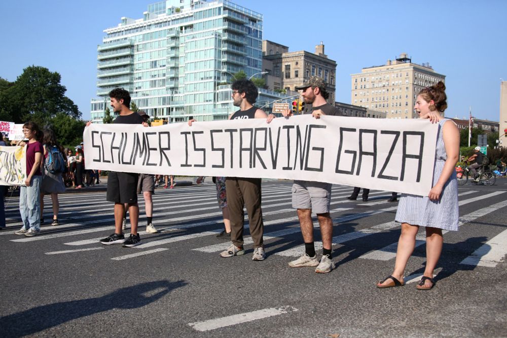 Four protestors holding a banner reading "SCHUMER IS STARVING GAZA" block traffic at Grand Army Plaza.