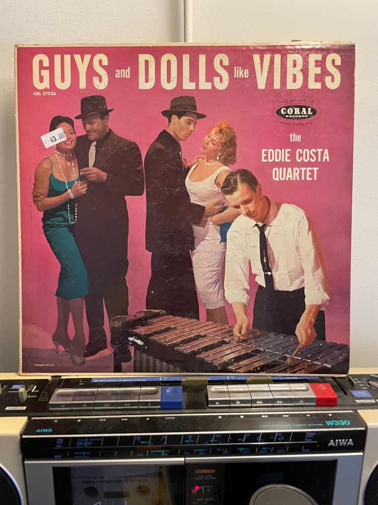 LP cover of an album titled GUYS AND DOLLS LIKE VIBES by the Eddie Costa Quartet sits atop a 1980s Aiwa boombox. The cover has a pink background and shows two couples who look like actors in production of Guys and Dolls smiling and holding each other close (maybe dancing?) while a guy in a white shirt and skinny black tie plays the vibes in the foreground. VIBES, BABY.