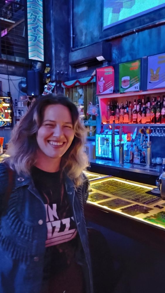 A super hot babe in a denim jacket and thin Lizzy T-shirt standing at a cyberpunk bar