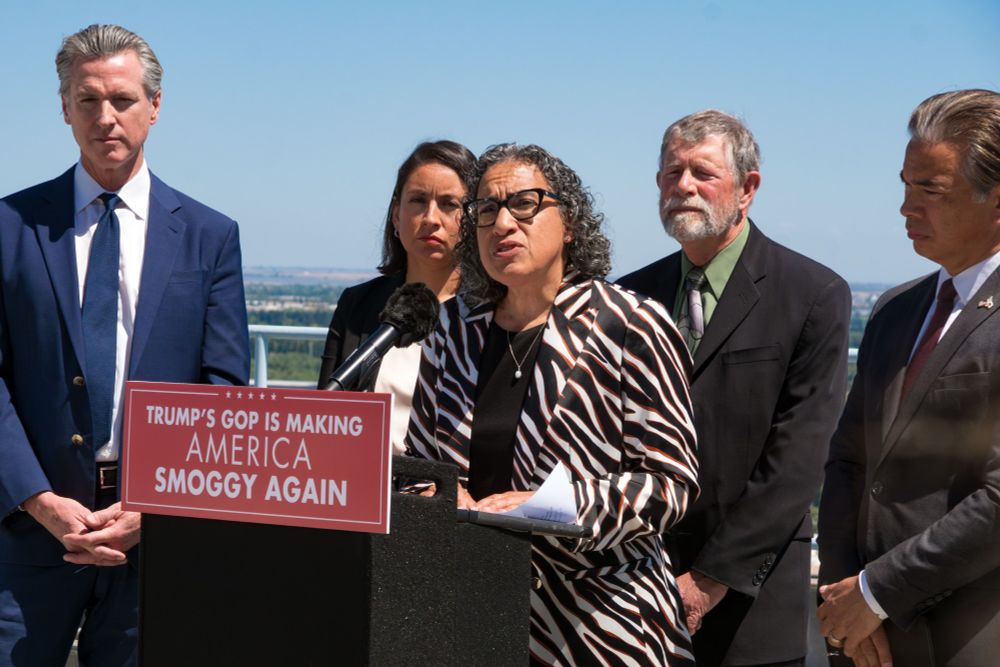 California officials stand together at a press conference. CARB Chair Liane Randolph speaks at a podium displaying a sign that reads, "Trump’s GOP is making America smoggy again." She is joined by Governor Gavin Newsom, Attorney General Rob Bonta, and other state leaders, addressing recent federal actions impacting clean air protections. A clear blue sky and cityscape are visible in the background. 