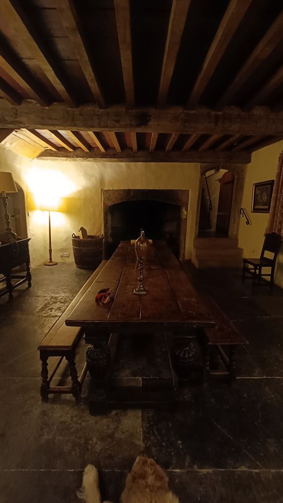 A medieval dining room with long oak table and benches, looking down the table towards an unlit stone fireplace with wood floor joists above and a flagstone floor.