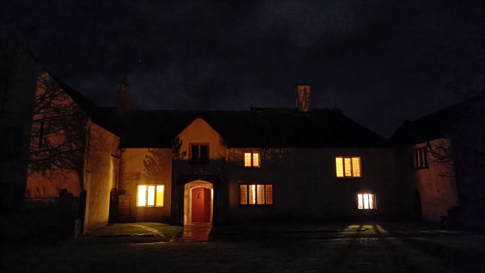 A very dark scene with an old house with windows glowing orange in the night, with a stone porch.