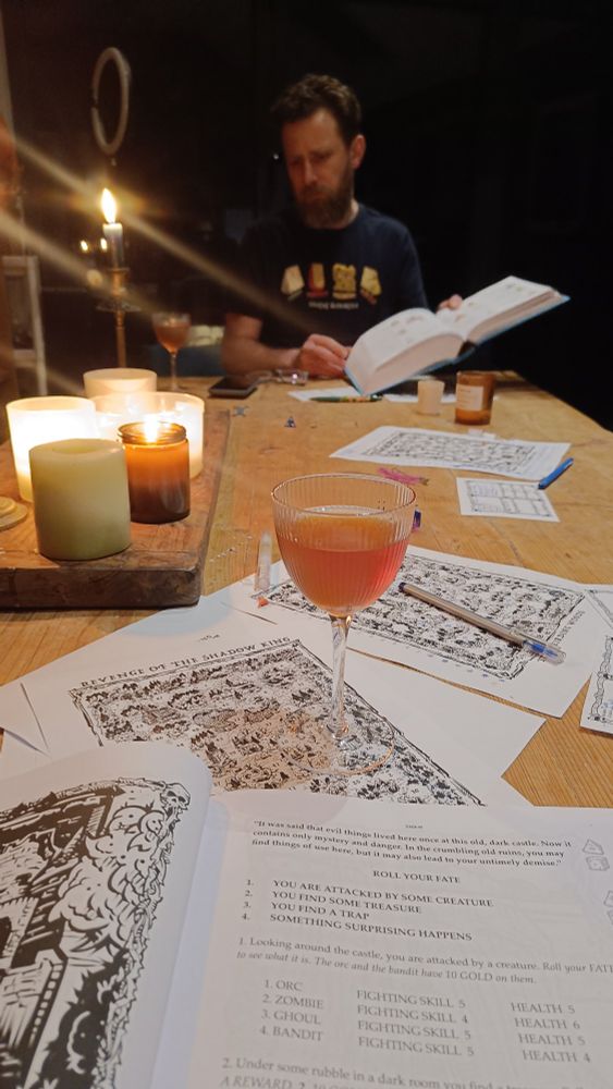 A table of candles and printed maps, an open book with quest info on it. A man in the background is looking through a big book. An orange cocktail in a small glass sits in the foreground.
