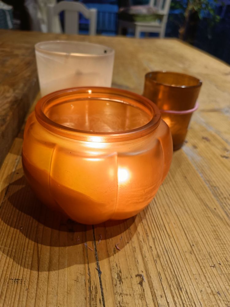 An orange pumpkin-shaped candle on a wooden table, two other lit candles in the background 