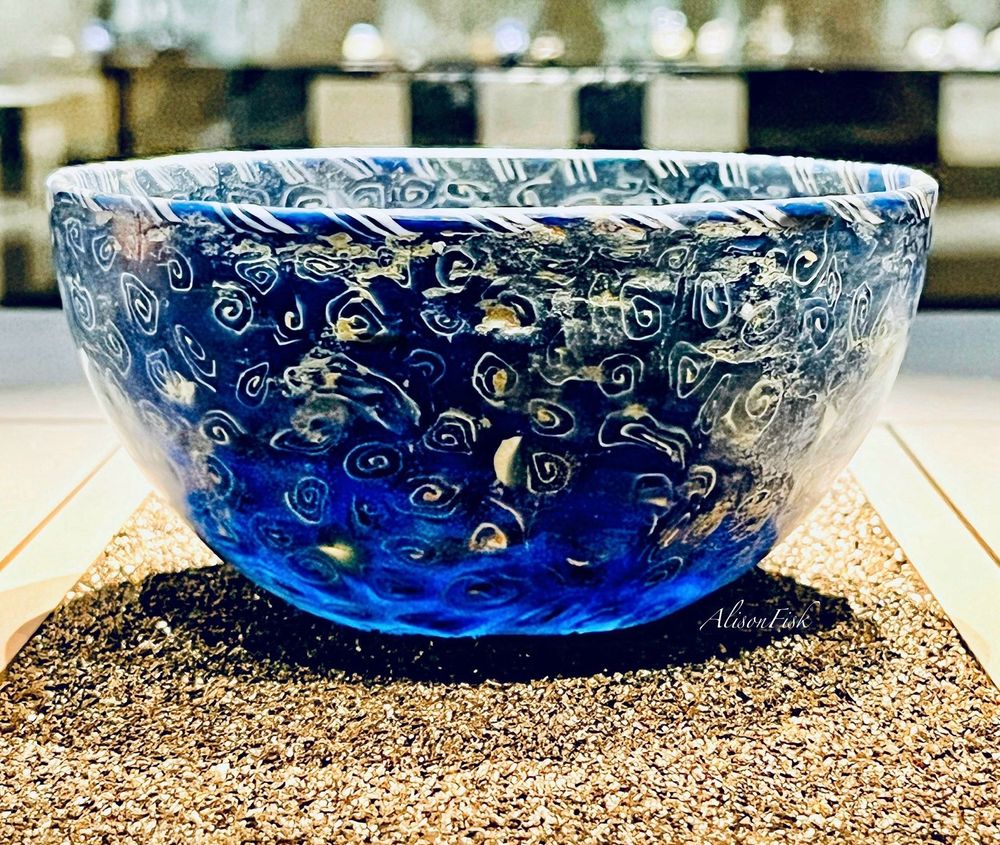 A hemispherical bowl made of mosaic cast glass. Translucent dark blue with opaque white spirals. There is slight corrosion and weathering of the glass. The bowl has been restored. Height: 7.2 cm, diameter: 13.1 cm. 

The effect of the museum lighting shining through the blue glass with white swirls brings to mind the painting ‘Starry Night’ by Vincent Van Gogh. 