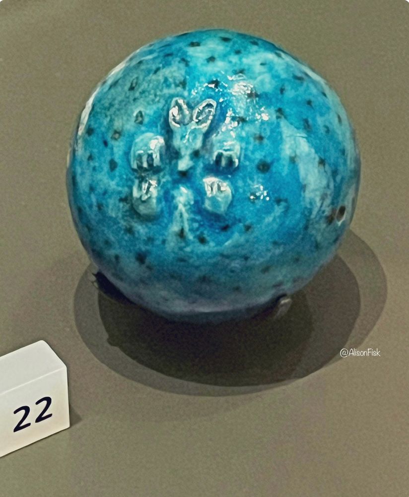 A blue spherical bead in the form of a hedgehog curled up in a defensive ball. Made of Egyptian faience, a ceramic material with a shiny blue glaze. The hedgehog’s face, ears, feet and tail are shown in relief, and the quills are shown by black dots painted over the surface. A hole for threading runs through the sphere