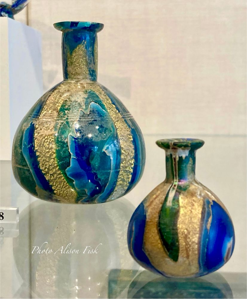My photo shows two Roman gold-band mosaic glass bottles on display at the Met Museum. Made of translucent cobalt blue, turquoise green, opaque white, and colorless glass encasing gold leaf. Both bottles have globular bodies and cyclindrical necks with horizontal everted rims. The larger bottle on the left measures 10.5cm in height and has a slightly concave bottom. The smaller bottle on the right measures 4.8cm in height and has a flat bottom.

According to the museum label: “Gold-band cast glass was a particularly opulent type of early Roman glassware. It combines canes of brightly colored translucent and opaque glass with strips of gold leaf encased between layers of colorless glass. Only a limited number of vessel shapes were made in this way, and some of the most common are small globular or carinated bottles such as these. They were presumably used to store expensive cosmetics and perfumes”.