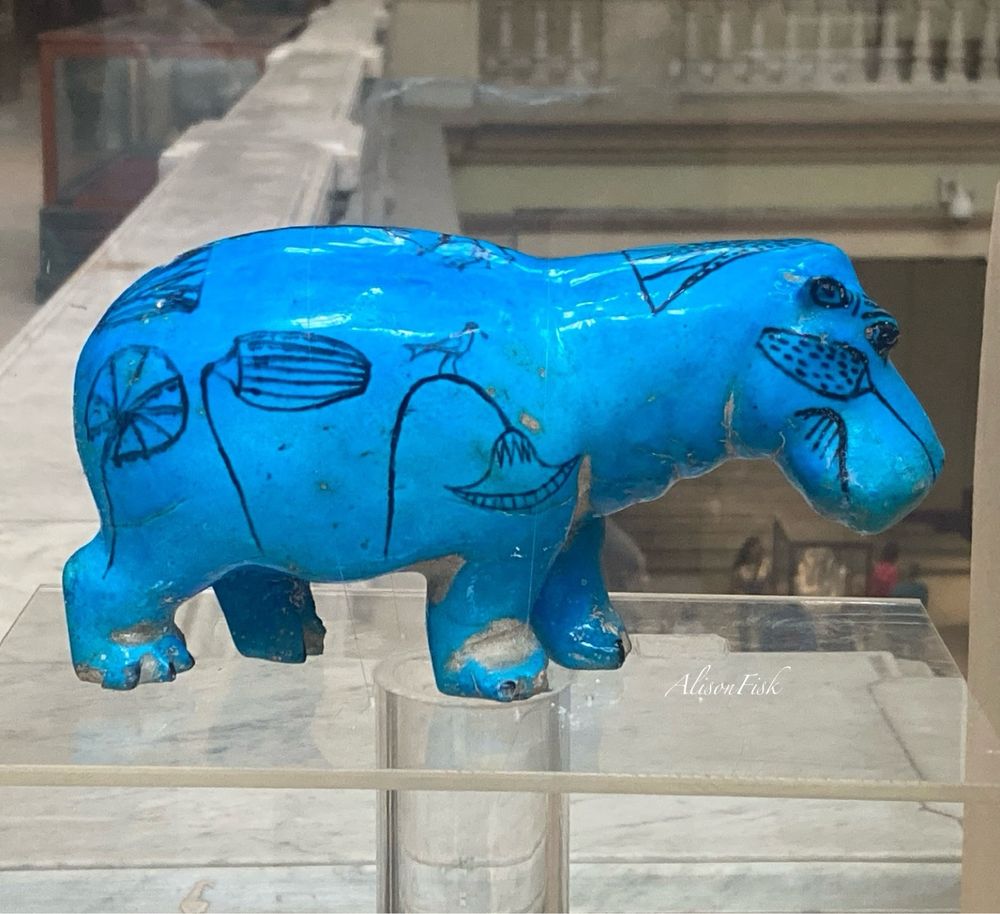 An ancient Egyptian statuette of a standing blue hippopotamus made of Egyptian faience, a ceramic material coated with bright blue glaze. The hippo is viewed from the side profile. It is decorated with black outlines of lotus plants. A little bird is perched on the stem of one lotus flower