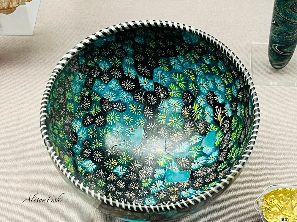 Hemispherical mosaic glass bowl viewed from above. It measures 13.2 cm in diameter and 7.5 cm in height. The glass is composed of sections of star patterned cane in blue, purple (looks black in my photo), and green, which appear as a floral pattern. The bowl has a black and white spiral rim. Made in the eastern Mediterranean about 200-100 BC. The bowl has been reconstructed and blue glass fragments which do not belong to this vessel have been used to fill missing areas.