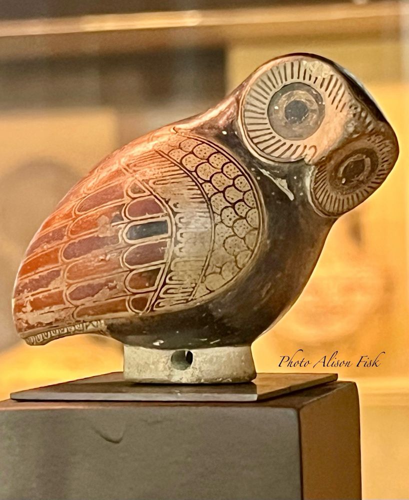 My photo shows a small Proto-Corinthian clay aryballos (perfumed-oil container) in the shape of an owl with a pouring hole in the tail. Height 5 cm, Width 6.4 cm. The owl is perched with its tail to the left and head to the right. It leans slightly forwards with head turned so that its large eyes face the viewer. The owl’s chest is painted black. The eyes and plummage are intricately detailed in black on a yellowy-cream slip. Three rows of feathers in the wing plumage are painted alternately in red and black. Dated c. 640 BC