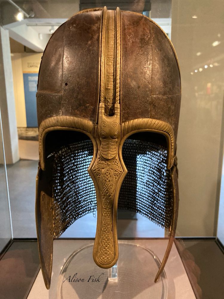 The Anglian York Helmet is composed of four main elements; a composite cap (brown in colour), two hinged cheek-pieces (also brown in colour) and a curtain of ring mail as a neckguard. The brass-coloured nose guard is covered with intricate interlaced animals. The 'eyebrows' have animal-head terminals, and an inscribed copper alloy band that runs over and across the crest of the helmet. The interlacing animals on the nose guard have open lips, comma-shaped eyes and spiral hips. Between the ‘eyebrows' and facing downward, is a larger animal head with a rounded snout and comma-shaped eyes, like those on the nose guard. This animal has ears on either side of its head that merge with the inscribed copper alloy band. An inscription on the band reads:
'IN. NOMINE. DNI. NOSTRI. IHV. SCS. SPS. D.
ET. OMNIBUS. DECEMUS. AMEN. OSHERE. XPI.'
This abbreviated Latin inscription may be translated as 'In the name of our Lord Jesus, the Holy Spirit, God and with all we pray. Amen. Oshere. Christ.'