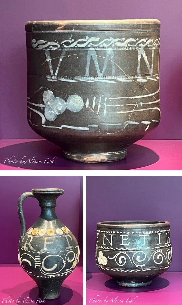 My photo collage shows three Roman black-slipped pottery vessels with white painted Latin inscriptions and white vine-like flowing decoration set against a rich pinky-purple display case background.

These so-called ‘motto beakers’, decorated with wine-themed Latin inscriptions painted in white slip, were produced in the Trier region during the mid 3rd century. They proved very popular and were exported to Britain and the Danube

Top image: cylindrical cup with straight sides with narrower base. The dark, almost black surface is decorated with large white letters V M N. The inscription around the cup reads VINIVM NOBIIS DA/ Give us wine! There is a band of rope-like white ornamentation near the rim. Beneath the lettering is a vinemotif with cluster of threeroundish ‘grape’ shapes. There are visible cracks where the cup has been restored. From Trier or surroundings, 280-315 AD.

Bottom left: elegant dark jug with a narrow base, rounded body, and long neck that flares outward to a broad lip. A single curved handle connects the neck to the upper body. The decoration includes the large white letters R E P and swirling white scroll ornamentation. The full inscription encircling the jug reads REPLE ME - Fill me up!  From Trier, St. Matthias, 280-315 AD.

Bottom right: A cylindrical beaker with a narrower base. White lettering near the top reads E N E T I B. The full inscription around the beaker reads B[ENIE T[BI S]|[T] - Well be to you! Beneath the text is a continuous vine-scroll design with stylised leaves or grapes. From Trier, Louis-Lintz-Straße, 260-270 AD.