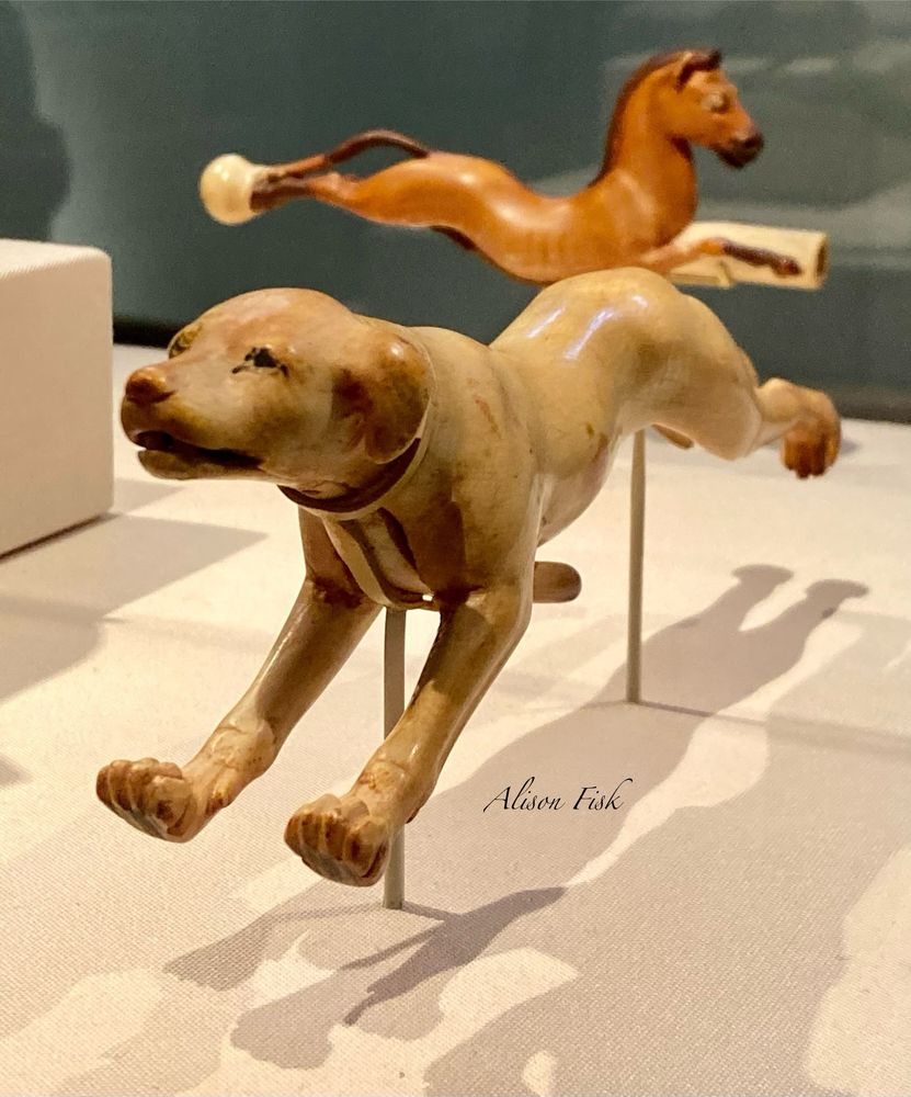 Ancient Egyptian ivory sculpture of a leaping dog. The ivory is now biscuit coloured. Dimensions: 18.2 cm  (L) × 6.1 cm (H) × 3.6 cm (W) A lever under the dog’s chest opens and closes the mouth. The inside of the mouth is red. The eyes are outlined in black. Dated Late Dynasty I8, I400-I350 BC.