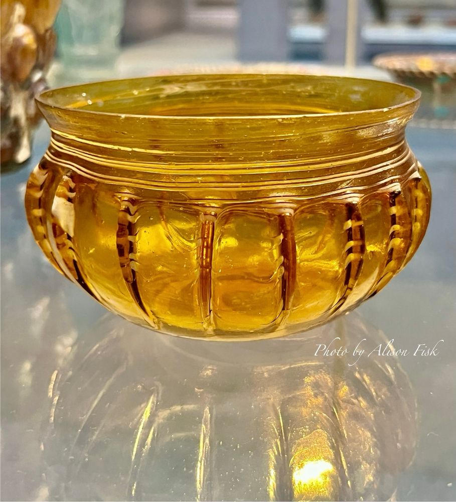 My photo shows a translucent amber coloured glass bowl with vertical rib decoration around the body with marvered white glass trails. The glass bowl glows in the display lighting as if it contains sunshine which reflects on the surface of the display case. Dimensions: Diameter 9.50 cm x Height 6 cm. On display at the British Museum.
