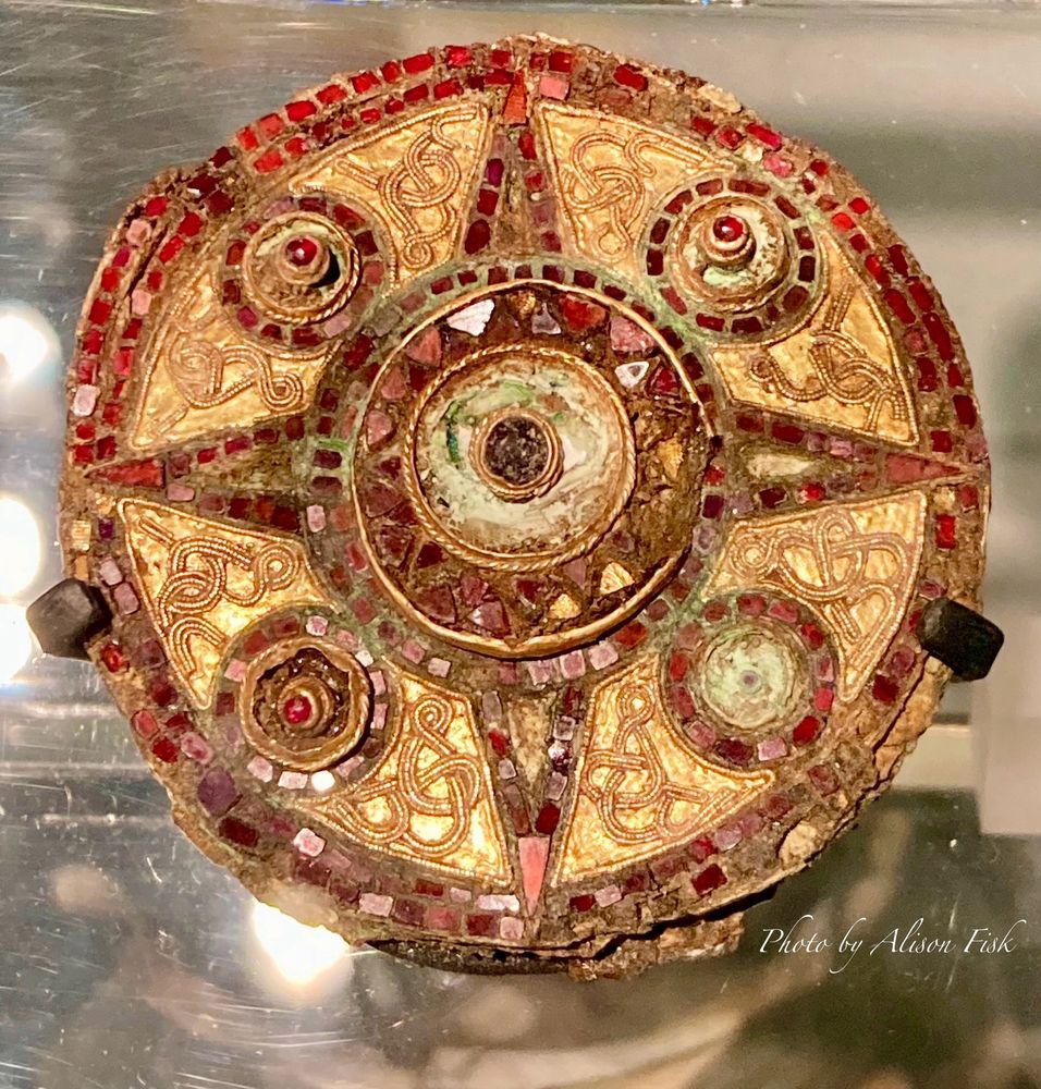 My photo shows a Saxon disc brooch made of copper embellished with gold plates and gold wire, set with five garnets in gold bosses, four of which are arranged around a larger central boss, and a mosaic of tiny polished garnet slabs around the outer edge of the brooch, and forming a four pointed star-like geometric shape around the central boss. The brooch measures 7 cm in diameter.

Excavated from a woman’s grave by AOC Archaeology in 2000. This type of brooch was a fashionable status piece amongst aristocratic Anglo-Saxon women in the AD 600s. The woman’s grave also contained glass beads and silver rings. 