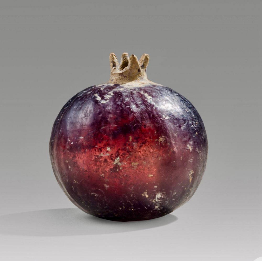 A small purple glass vessel in the shape of a pomegranate probably used as a container for perfumed oil. Height 6.9 cm (2.7 in), Diameter 6.6 cm (2.5 in) The lighting enhances the translucent purple glass which replicates the colour of a ripe fruit. There is some encrustation particularly around the calyx which has one lobe missing.