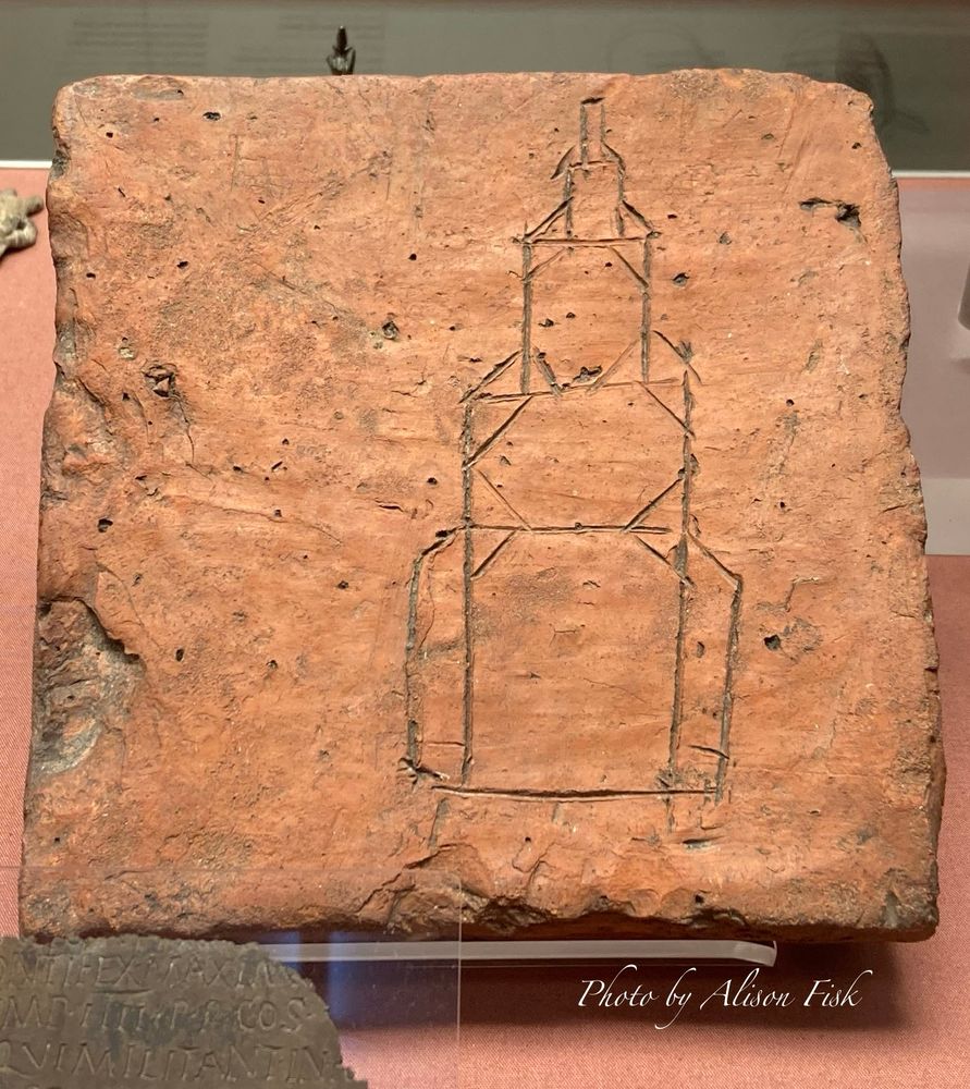 My photo shows a square Roman floor tile made of red clay. Inscribed into the clay is a drawing of what appears to be a Roman lighthouse (faros). The lighthouse has four storeys, which are stepped inwards at each level. Tile width 20 cm. Two Roman lighthouses flanked the harbour at Dover, the main base of the Roman fleet ‘Classis Britannica’. On display at the British Museum. 
