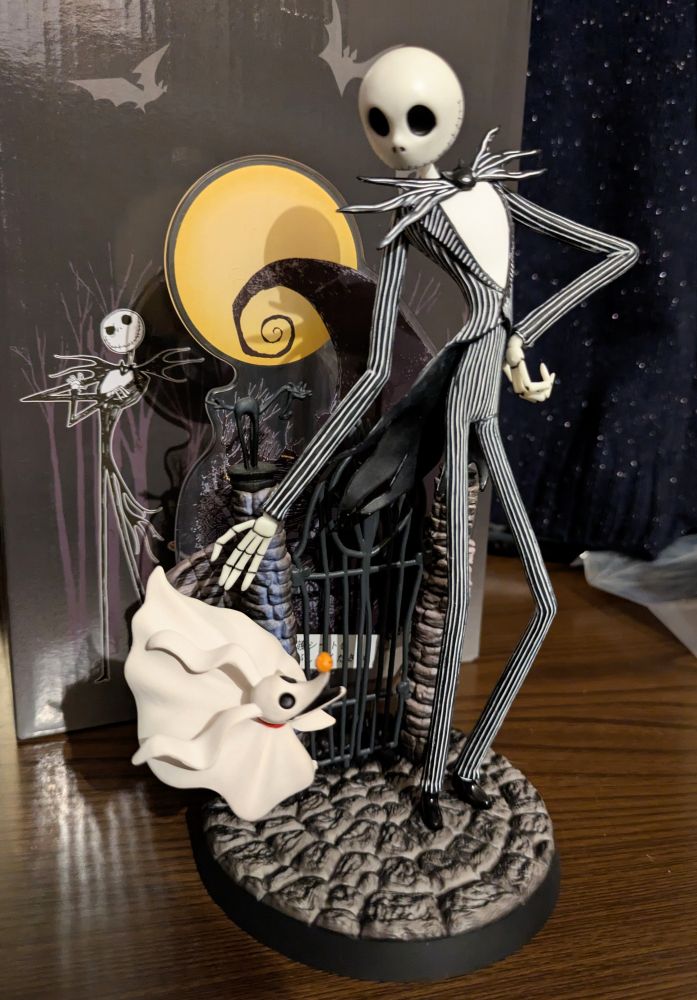Photo of a figurine of Jack Skellington and his ghost dog Zero from "The Nightmare Before Christmas", standing in front of a gate. Behind the figurine is the box it came in.