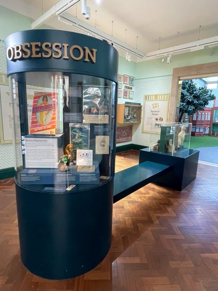 A small museum gallery with dark wooden parquet floors. A central blue cylindrical display case is topped with gold letters spelling “obsession”. Inside the case are Egyptomania Museum objects. Other cases and panels are on the walls around the gallery.