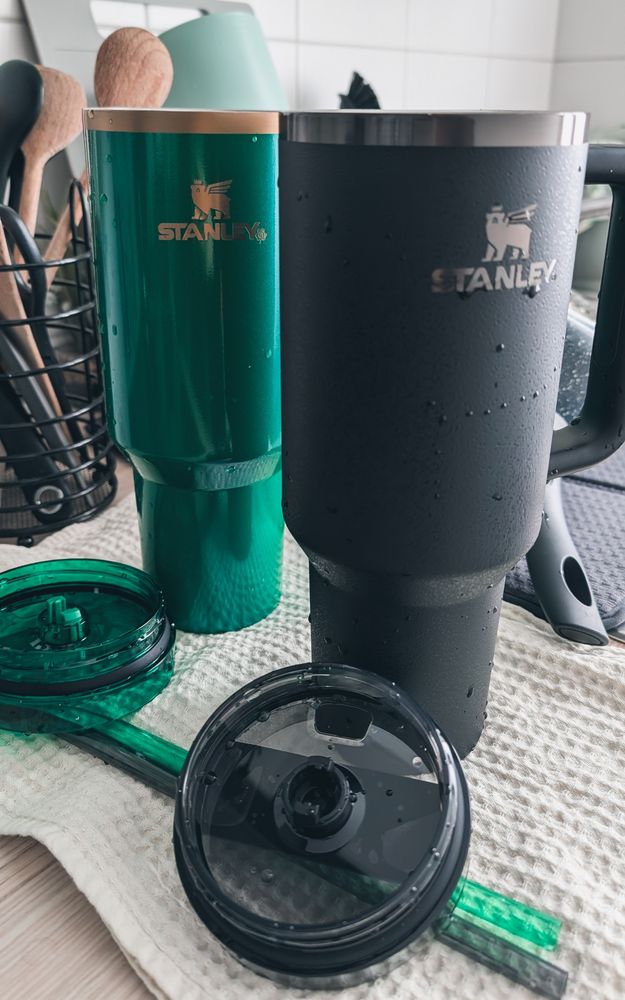 Zwei Stanley-Thermobecher, einer grün und einer schwarz, stehen nebeneinander auf einem Küchentuch. Ihre Deckel und Trinkhalme liegen im Vordergrund, noch nass vom Spülen.