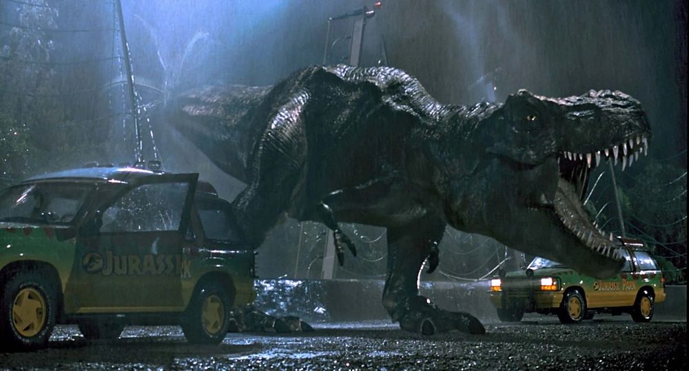 Still photo from Jurassic Park. The T-Rex, having crashed through the exhibit fence, stands on the road and roars in the rain.
