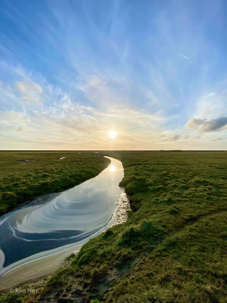 Ein Wassergraben verläuft durch Wiesen, die Sonne scheint und spiegelt sich im Wasser wider. Am Horizont des blauen Himmels befindet sich ein Wolkenschleier. 