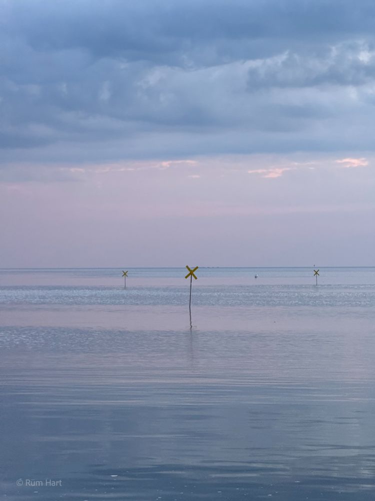 Die glatte Nordsee am Abend. Der Himmel schimmert in einem rosafarbenen Ton, während sich darüber eine ausgedehnte graue Wolke erstreckt. Es stehen drei Kreuze im Wasser, die den Badebereich markieren. Der Himmel spiegelt sich im Wasser wider. 