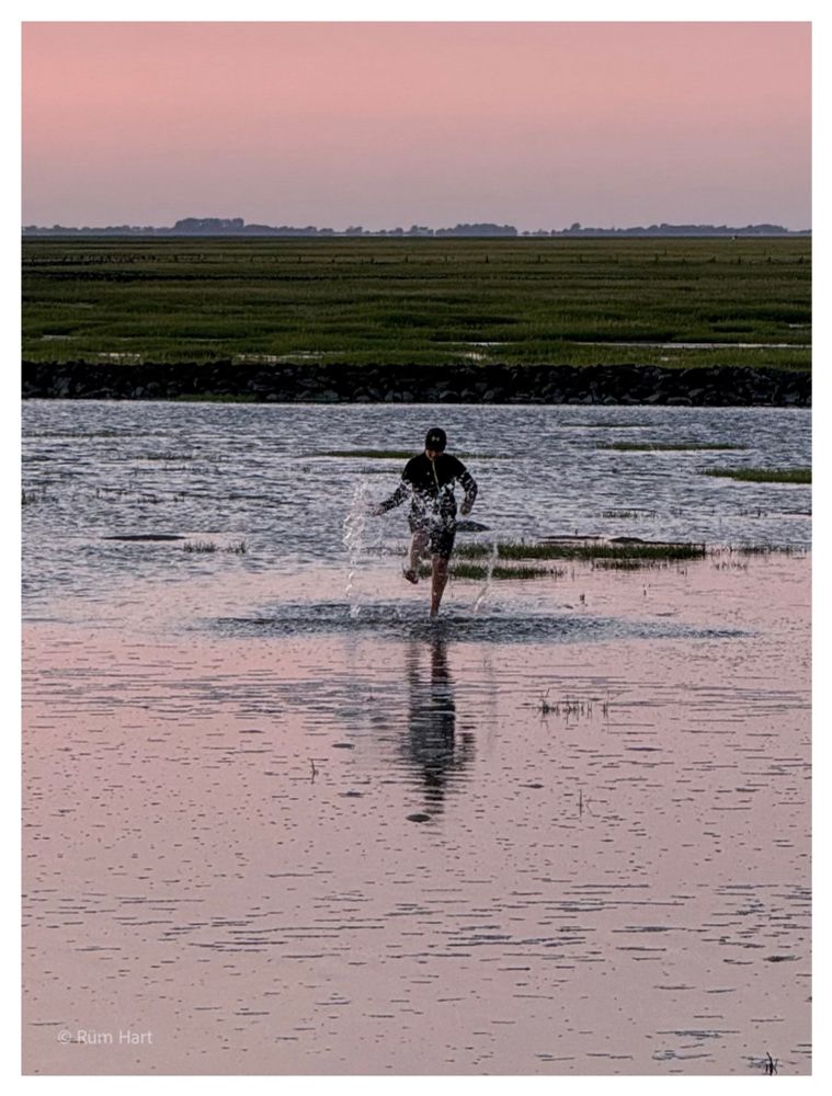 Ein Mann steht im Meer und schleudert mit seinen Füßen Wasser nach oben. Im Hintergrund verläuft eine Buhne und dahinter beginnen die Salzwiesen. In weiter Ferne befindet sich eine Baumreihe. Das Wasser spiegelt den rosafarbenen Himmel wider. 