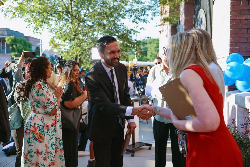 Zohran smiling and shaking hands with staff outside a school.
