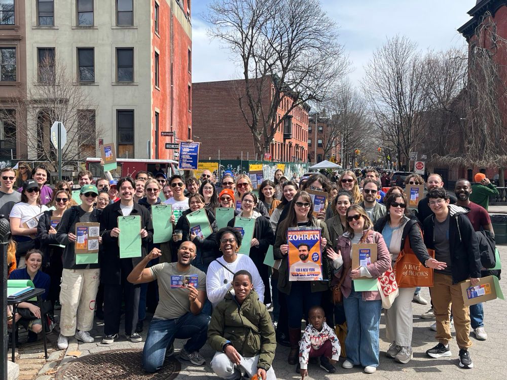 State Senator Jabari Brisport and Assemblymember Phara Souffrant Forrest in the front row of a huge crowd of volunteers posing with clipboards and Zohran posters on a sunny day in Fort Greene Park.