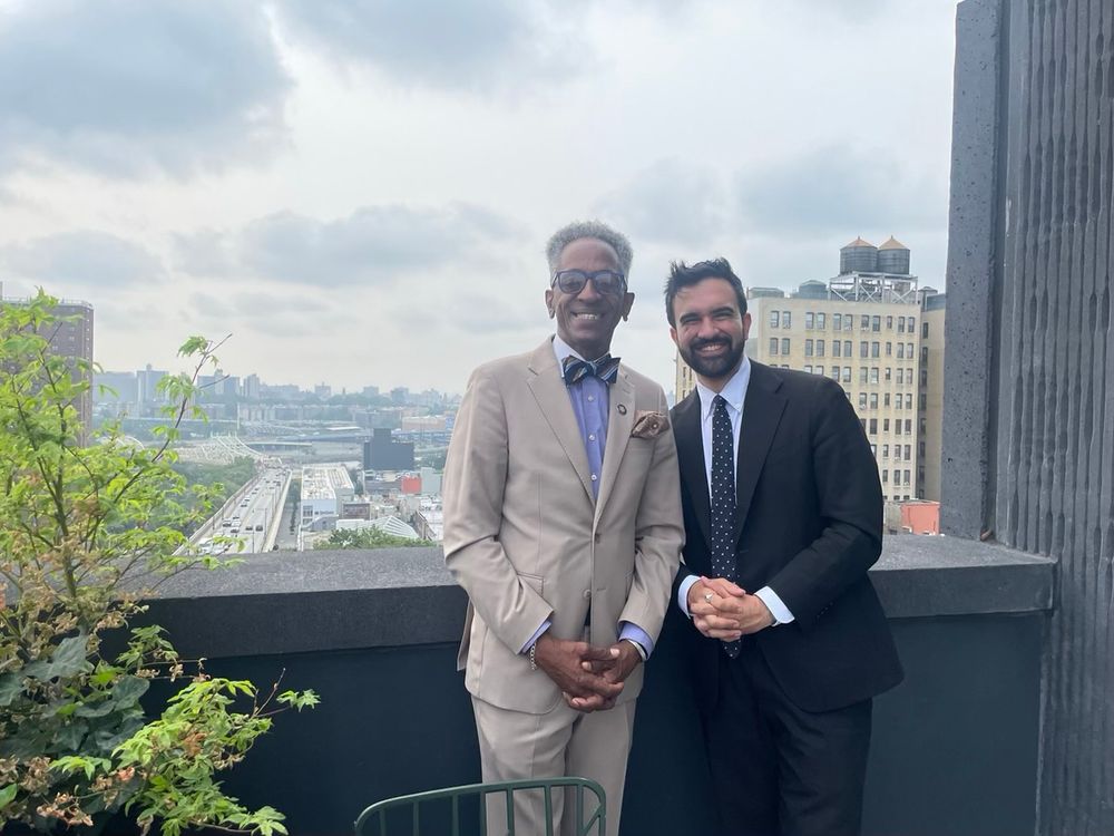 Zohran and Al Taylor standing together facing the camera on a balcony of a high-rise building. 