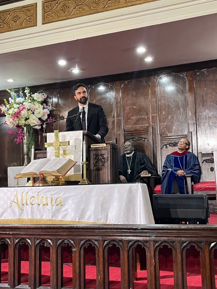 Zohran standing at an altar speaking with two men in robes sitting behind him. 