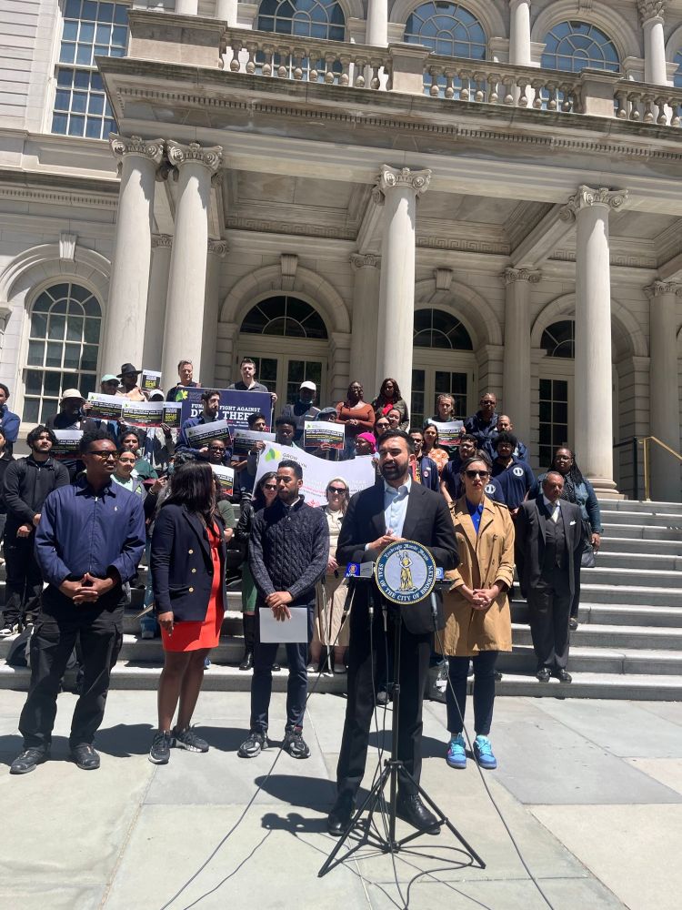 Zohran at a podium in front of City Hall, with elected officials standing behind him. 