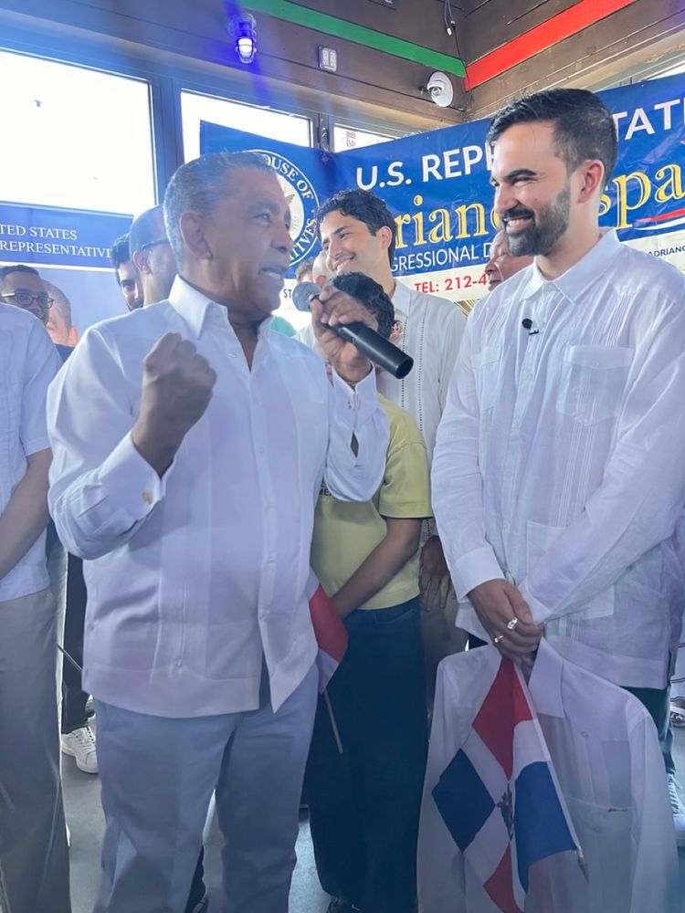 Congressman Adriano Espaillat speaks into a microphone at his annual Dominican Day Parade Breakfast. Zohran stands next to him smiling.