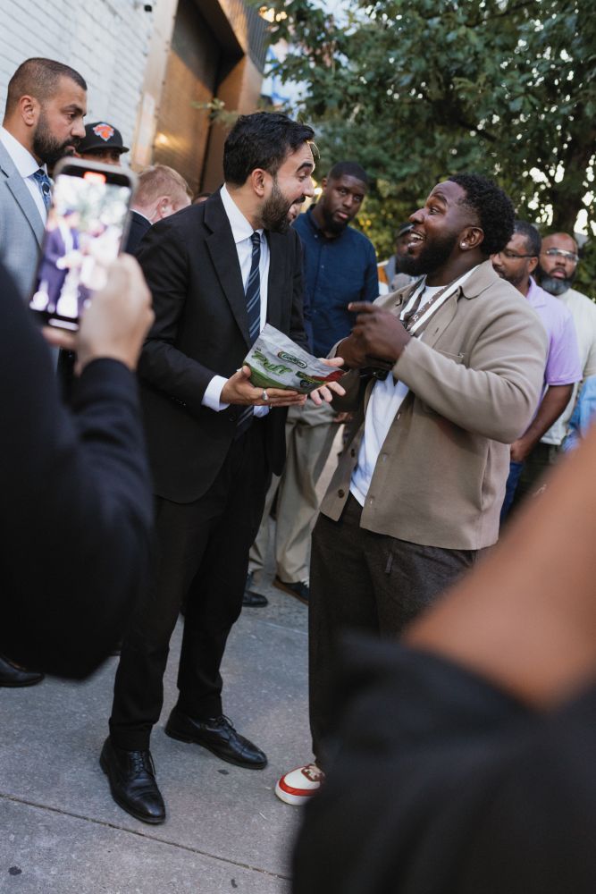 Zohran smiling with an attendee on the sidewalk outside the event. 