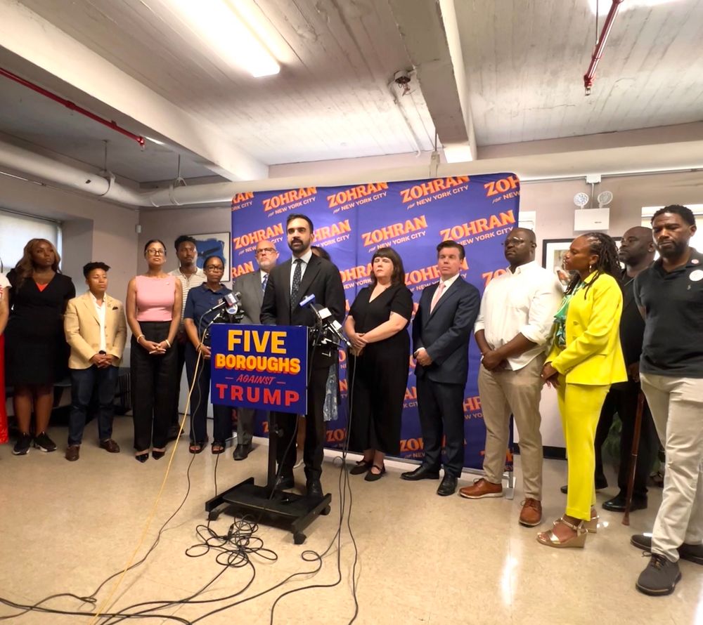 Zohran at a podium in front of a group of Brooklyn elected officials. The podium sign says “Five Boroughs Against Trump.”