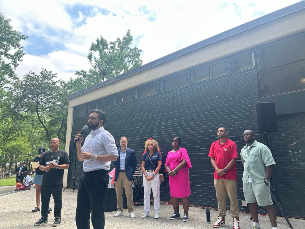 Zohran holding a mic addressing a crowd at a park with elected official standing behind him.