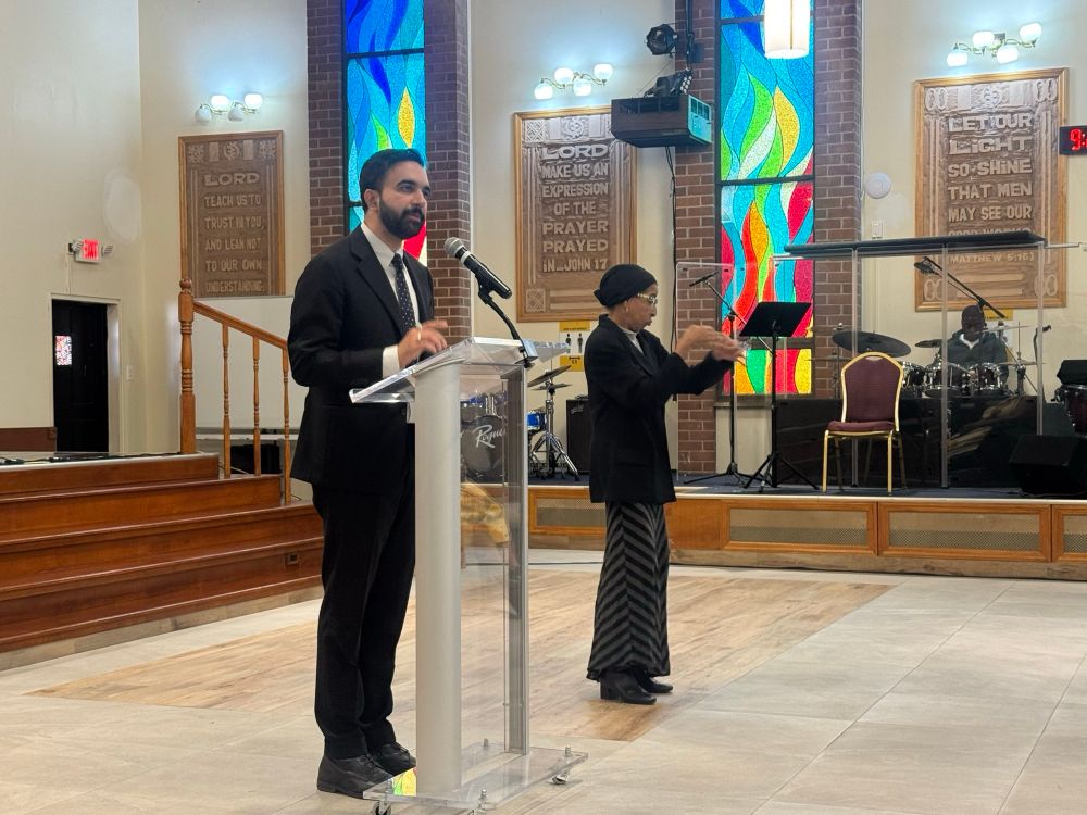 Zohran standing at a podium in a church with beautiful stained glass windows.