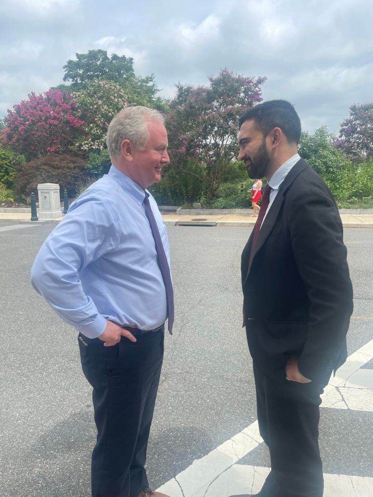 Senator Chris Van Hollen, hands on hips, stands facing Zohran Mamdani. Both are smiling. 