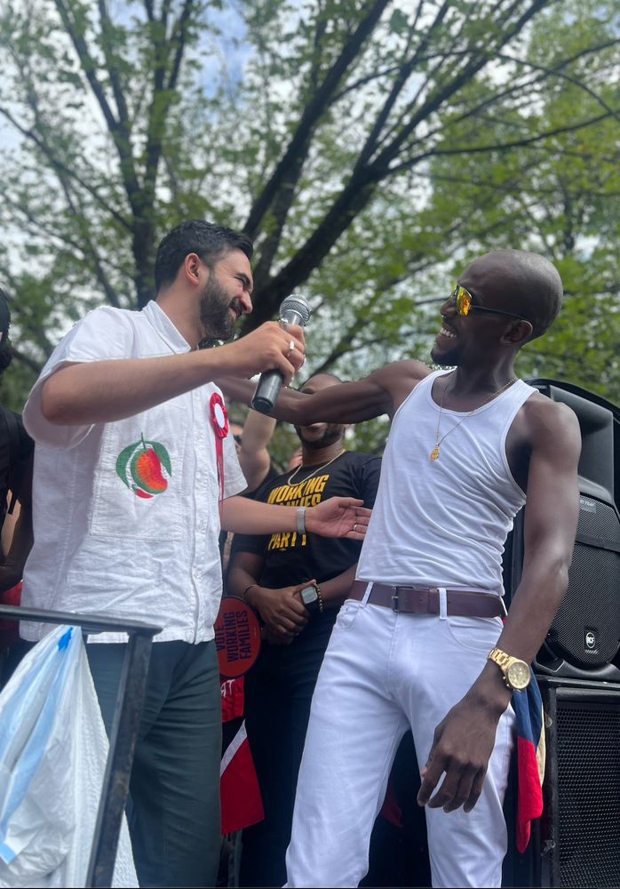 Zohran about to embrace a man in a white tank top on a parade float. Both are smiling. 