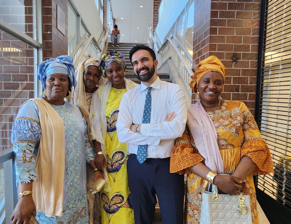 Zohran stands, arms folded and smiling, with a group of Bronx women at the Guinean community event in Co-op City. 