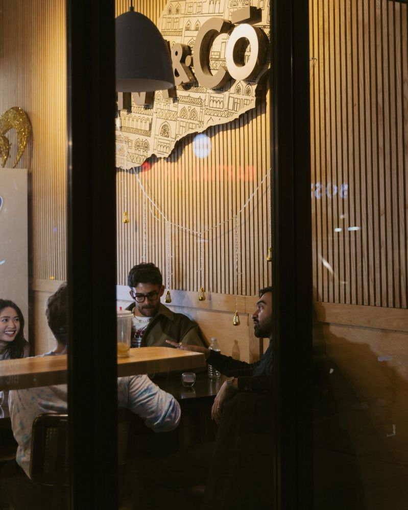 A photo taken from outside Moka & Co. looking in at Zohran and Hasan seated at a table chatting.